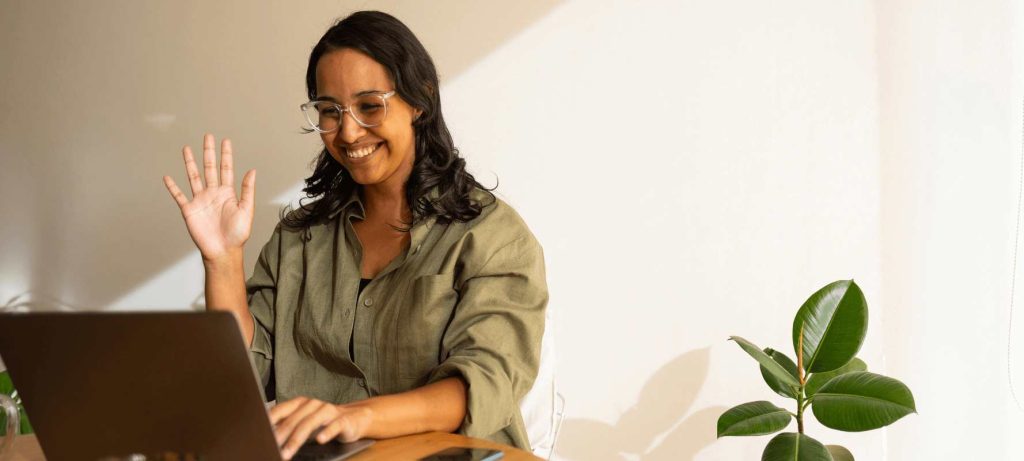 A smiling woman sitting at a table, waving toward here laptop screen.