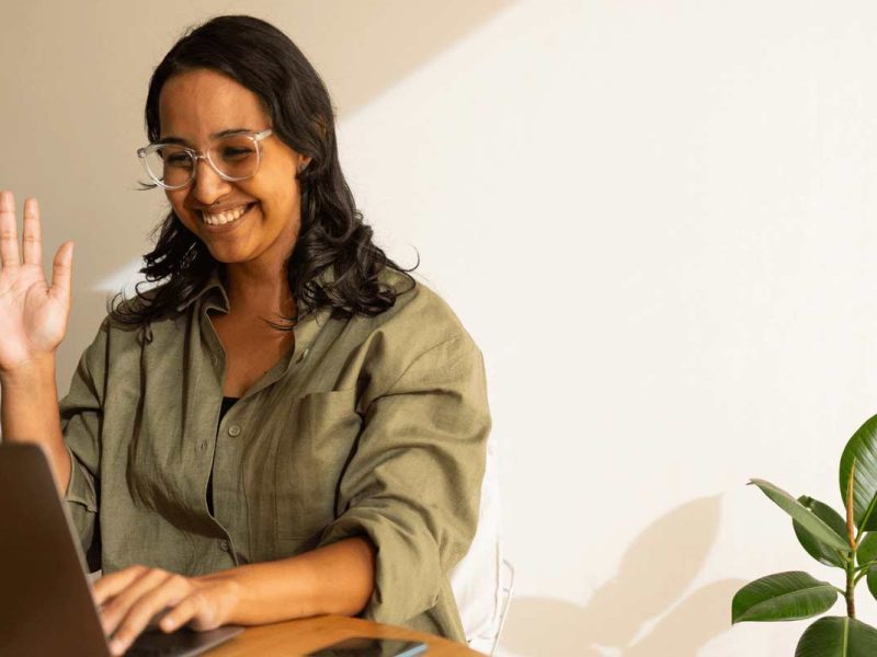 A smiling woman sitting at a table, waving toward here laptop screen.
